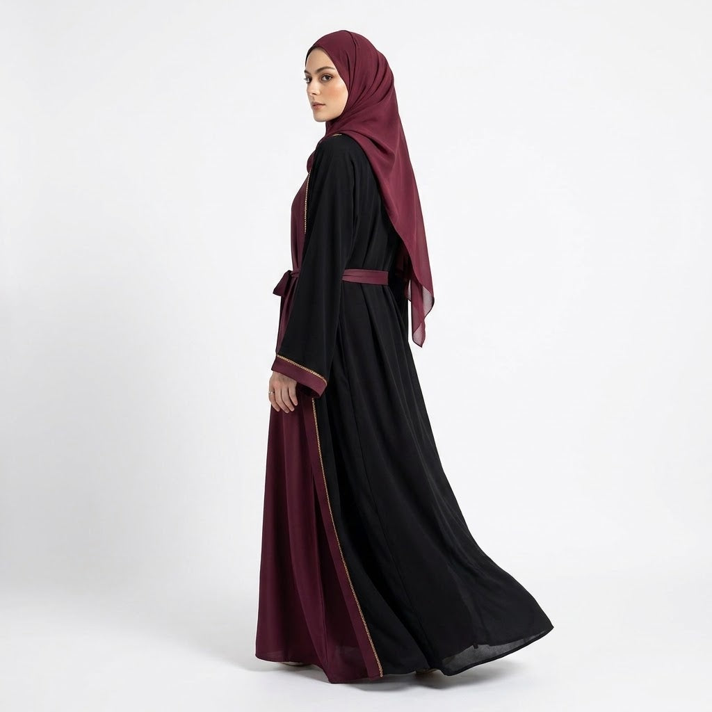 A woman wearing a long black abaya with deep burgundy trim and a matching headscarf, shown in side profile against a plain light gray studio background.