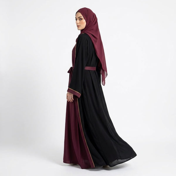 A woman wearing a long black abaya with deep burgundy trim and a matching headscarf, shown in side profile against a plain light gray studio background.