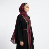 A woman wearing a black abaya with deep burgundy trim and a matching headscarf, shown in profile against a plain light gray studio background.