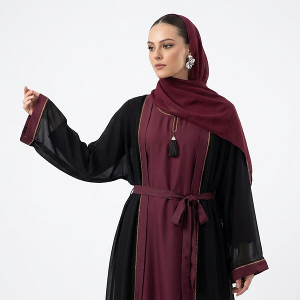 A woman modeling a traditional modest outfit featuring a black long-sleeved tunic with sheer sleeves and a deep burgundy waist sash, paired with a matching burgundy hijab, against a plain white studio background.
