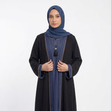 A woman modeling a black abaya with blue trim and a matching blue hijab, standing against a plain light gray studio background.