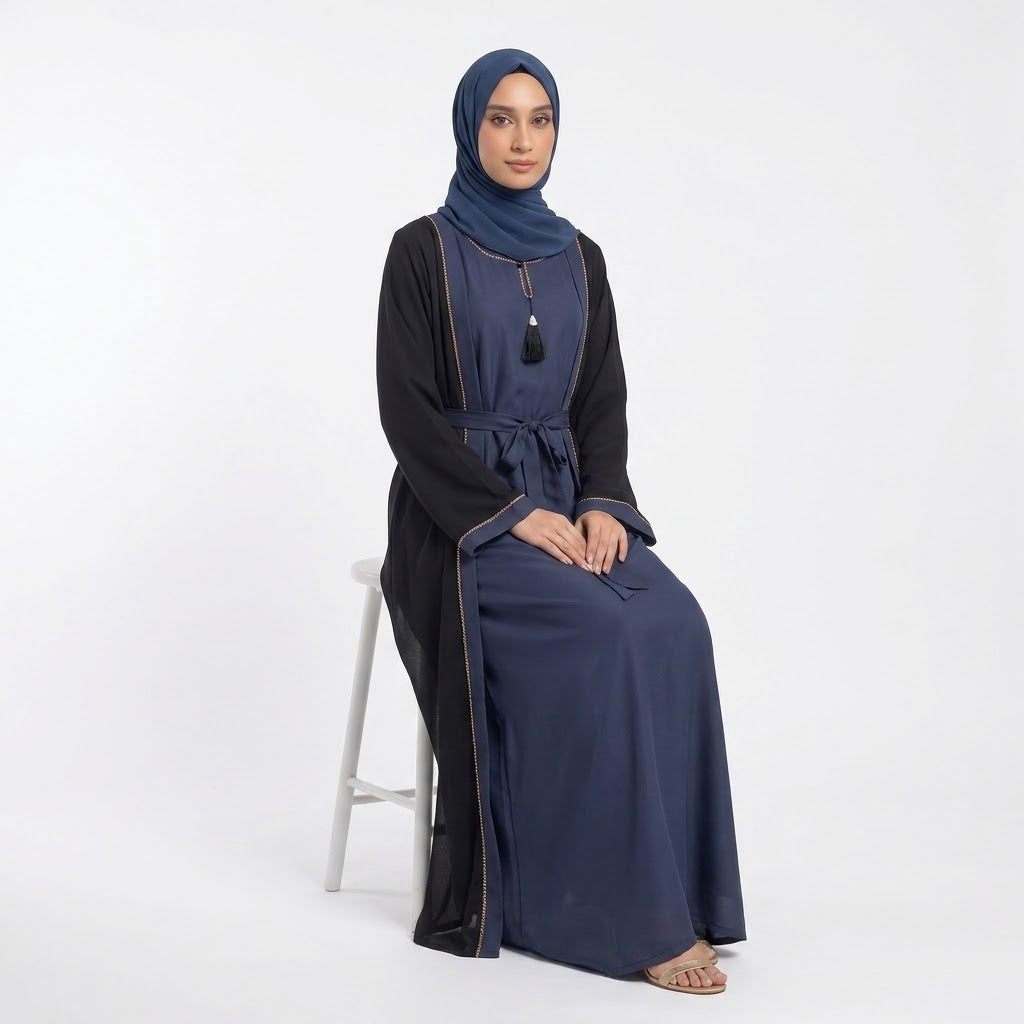 A woman wearing a navy blue abaya with black sleeves and a matching navy hijab, seated on a white stool against a plain white studio background.