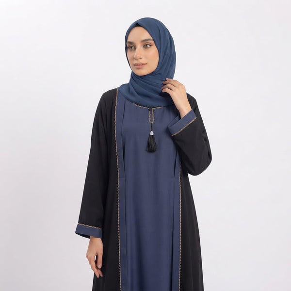 A woman wearing a navy blue abaya with black sleeves and a matching navy hijab, standing against a light gray studio background.